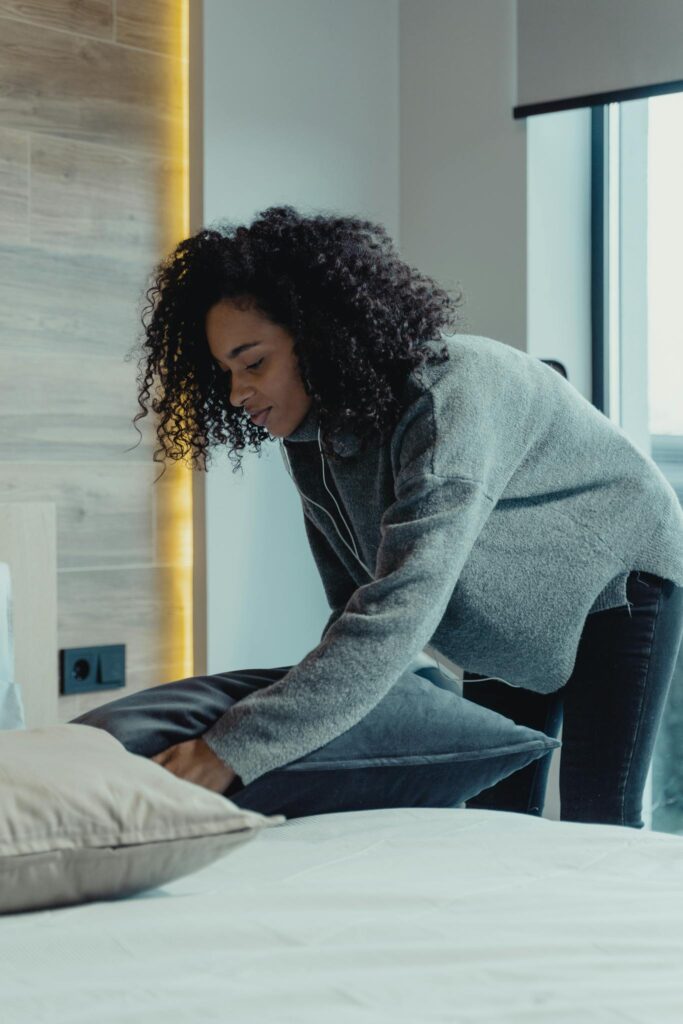 A woman making a bed in a modern bedroom with natural light, reflecting a cozy and tidy home environment.