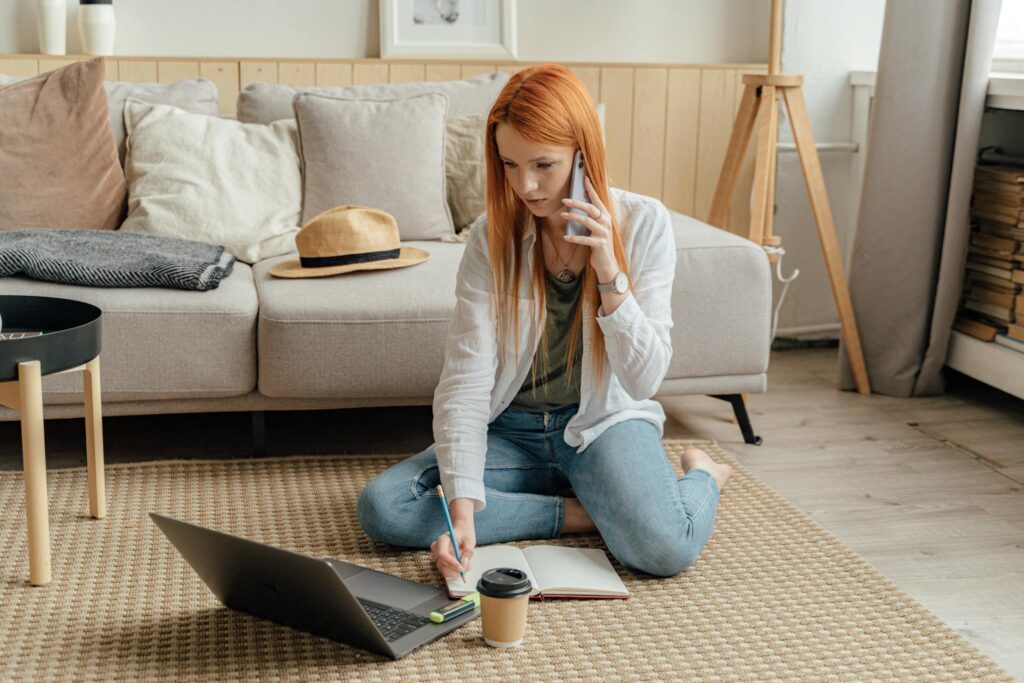A woman sitting on the floor using a laptop and phone in a modern home office environment.